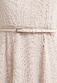 Robe en dentelle blanche avec des motifs complexes, cintrée à la taille par une fine ceinture crème avec une petite boucle.