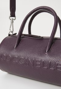 Sac à main en cuir violet à textures en relief, de forme ronde, avec deux poignées supérieures, logo "COCCINELLE" en relief et bandoulière amovible.