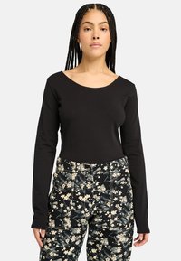 Schwarzes Langarmoberteil mit weitem Ausschnitt, kombiniert mit hoch taillierten Hosen mit dunklem Blumenmuster, die Taschen und einen strukturierten Stoff aufweisen.