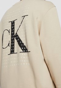 Sweatshirt beige avec un logo noir "CK" bien visible. L'arrière comporte un texte répétitif en blanc : "Calvin Klein Jeans Established 1978." Texture douce.