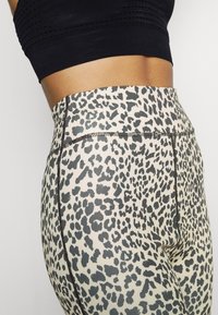 Leggings taille haute en tissu extensible, avec une base beige ornée d'un motif léopard noir et des détails de couture visibles.