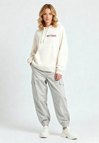 Felpa crema con scritta rossa, abbinata a pantaloni cargo grigi chiari con tasche laterali e polsini elastici. Sneakers bianche completano il look.