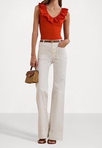 Červený bezrukávový body s volánkovým lemem, bílé kalhoty s širokými nohavicemi, předními kapsami a zapínáním na knoflík, a malá tkaná kabelka.