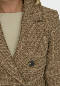 Blazer double boutonné en tweed marron avec un tissu texturé et des détails de boutons noirs proéminents. La couche intérieure présente un tissu de couleur claire.