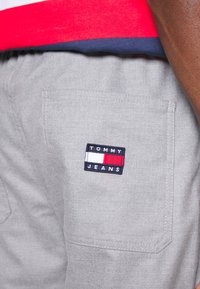 Šedé šortky s texturovanou tkaninou mají černou nášivku s nápisem "TOMMY JEANS" a červenými a bílými akcenty. Viditelné standardní kapsy.