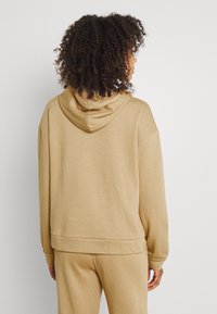 Sudadera con capucha color marrón claro, de textura suave, corte holgado y puños acanalados. El diseño cuenta con un bolsillo canguro y un dobladillo redondeado en la cintura.