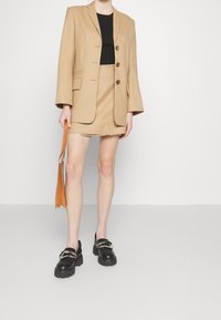 Blazer beige à manches larges avec trois boutons, assorti à une mini-jupe assortie. Haut noir et mocassins noirs épais, tenant un sac orange.