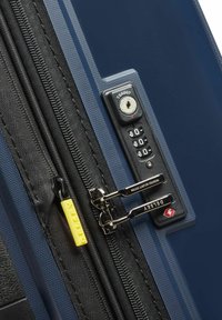 Maleta rígida azul en primer plano que presenta un zipper negro con un tirador amarillo, candado de combinación y herrajes de acero pulido.