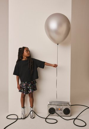 Młoda dziewczyna w czarnej koszulce i błyszczącej spódnicy trzyma srebrny balon przywiązany czarnymi sznurkami do retro boomboxa na beżowym tle.