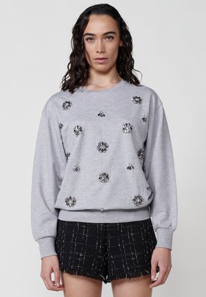 Sudadera gris con adornos de lentejuelas plateadas en patrones florales. Presenta mangas largas y un corte relajado, combinada con pantalones cortos negros texturizados.