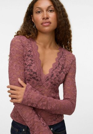 Femme aux cheveux bouclés portant un haut en dentelle mauve à manches longues avec un col en V festonné et un jean en denim foncé devant un fond uni.
