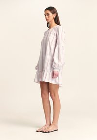 Donna che indossa un vestito bianco lungo e dal taglio ampio con strisce verticali multicolori e un orlo arricciato, abbinato a sandali piatti trasparenti.