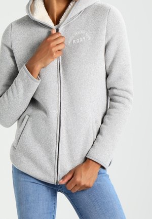 Kvinde iført lysegrå hættetrøje med lynlås og fleecefor, blå jeans, hånd nær lynlåsen og synlig lomme.