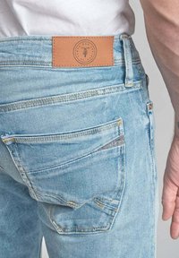 Jeans en denim bleu clair avec une patch en cuir à la taille, dotés de poches arrière standard et d'une texture délavée.