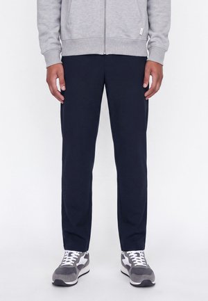 Personne portant un pantalon noir droit, un sweat gris zippé et des baskets grises et blanches, debout devant un fond blanc uni.