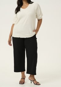 Blouse blanche texturée avec des manches courtes bouffantes, accompagnée de pantalons larges noirs et de sandales à talons noires, mettant en valeur la silhouette de la tenue.