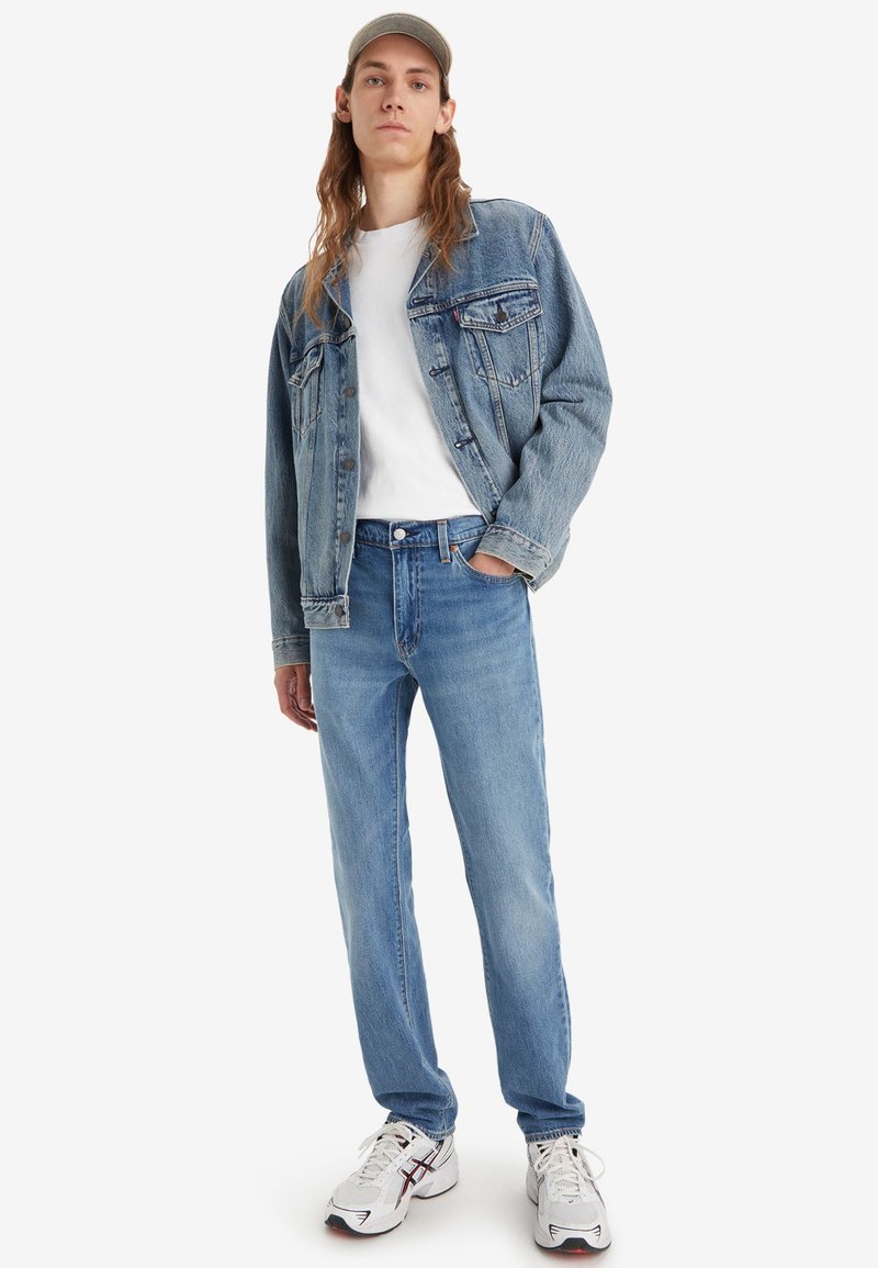 Joven vestido con una chaqueta de mezclilla clara, camiseta blanca, vaqueros azules, zapatillas blancas y una gorra beige, de pie con una mano en el bolsillo.