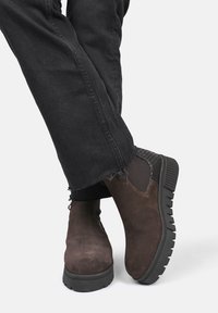 Bruine suede enkellaarzen met textuur zwarte zijpanelen, rubberen zool en een lage hak, gecombineerd met gescheurde zwarte jeans.
