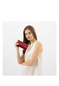 Portafoglio in pelle verniciata rossa con texture a coccodrillo, dotato di una chiusura in metallo argentato, tenuto da una persona vestita con una maglietta senza maniche di tonalità neutra.