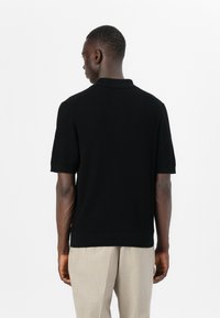 Μαύρο ριπ πόλο μπλ�ουζάκι κοντομάνικο με γιακά, με υφή. Συνδυασμένο με ανοιχτό μπεζ παντελόνι, σε θέα από πίσω.