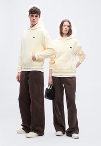 Δύο μοντέλα φορούν μπεζ φούτερ με κουκούλα και καφέ παντελόνια με φαρδύ πόδι, το ένα κρατά μια μικρή μαύρη τσάντα, στέκονται μπροστά σε λευκό φόντο.