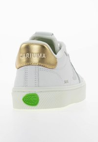 Białe sneakersy z metaliczną złotą zakładką na pięcie z napisem "CARIUMA" oraz zielonym logo na podeszwie, teksturowanym wierzchem i klasycznym wzorem sznurowania.