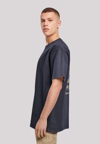 Námořnická modrá bavlněná tričko s volným střihem, krátkými rukávy a zlatým grafickým designem na zádech, předváděné na modelu z boku.