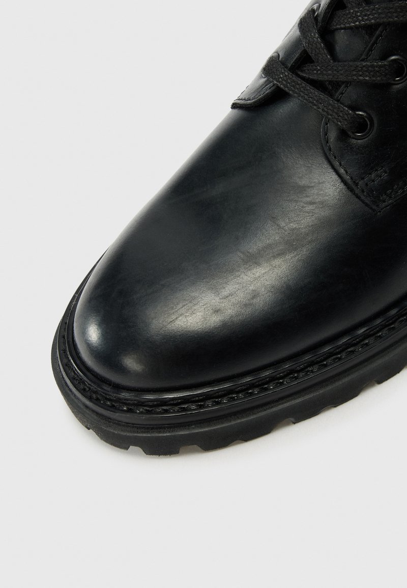Zapato de cuero negro con punta redondeada, parte superior texturizada y suela resistente con un detalle de costura a lo largo del borde.