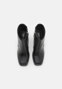 Botines de cuero negro hasta el tobillo con punta cuadrada, textura suave, cierre de cremallera lateral y elementos de metal plateado.