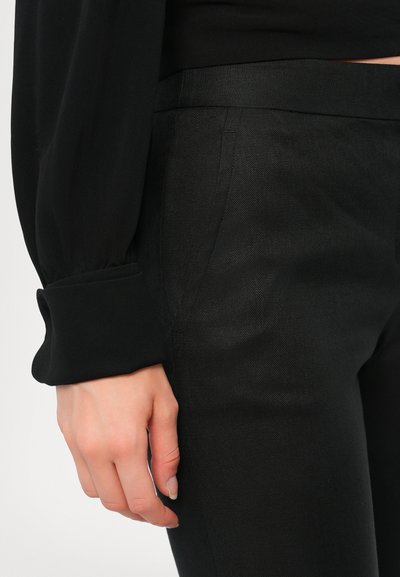 Pantalons noirs ajustés en tissu texturé, avec un devant plat, un léger brillant et des poches latérales, associés à un haut noir à manches longues.