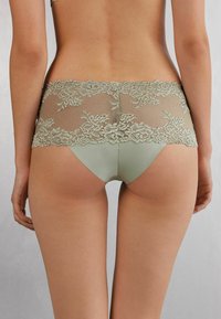 Ropa interior con espalda de encaje de tonalidad verdosa; presenta un detalle de encaje floral, un frente suave y satinado, y un diseño ajustado.