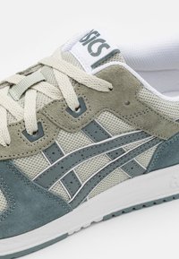 Light gray and green mesh and suede sneaker with beige laces, featuring a white tongue and gray logo, and layered design elements.