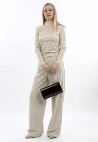 Top beige con paillettes, pantaloni high-waisted beige, che tiene una pochette nera con tracolla a catena, su uno sfondo bianco semplice.