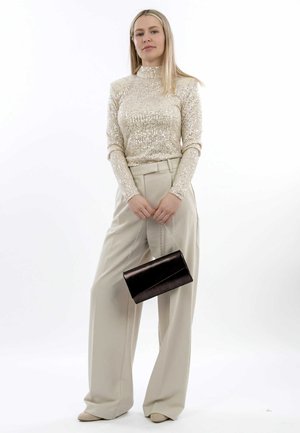 Haut beige pailleté, pantalon taille haute beige, tenant une pochette noire avec une chaîne, sur fond blanc uni.