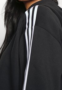 Svart sweatshirt med tre vertikala vita ränder på axeln, mjukt material och avslappnad passform.