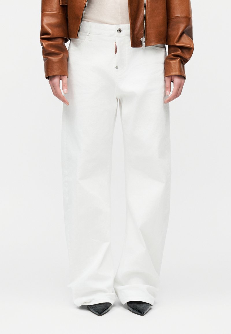 Άτομο που φοράει λευκό παντελόνι με φαρδύ πόδι, μαύρα μυτερά παπούτσια, καφέ δερμάτινο μπουφάν και λευκό τοπ, πάνω σε απλό φόντο.