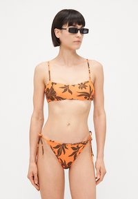 Oranžs bikini komplekts ar ziedu rakstiem, plāniem regulējamiem siksnām un sānu auklām apakšā. Gluda, pieguļoša auduma tekstūra.