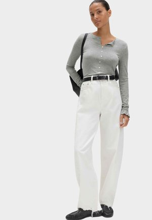 Frau trägt graues, langärmliges Top mit Knöpfen, hoch taillierte weiße Jeans mit schwarzem Gürtel, schwarze Schuhe und trägt eine schwarze Schultertasche.