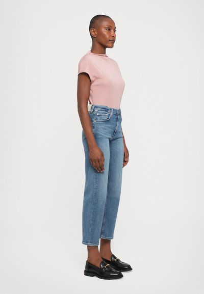 Woman standing sideways wearing a light pink short-sleeve top, high-waisted blue jeans, and black loafers on a plain background.
