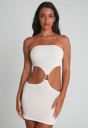 Vrouw met lang, donker haar, gekleed in een witte strapless gebreide jurk met uitsnijdingen en een metalen ringdetail bij de taille, staande tegen een effen achtergrond.