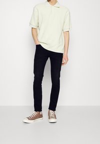 Hombre vestido con una camiseta polo de manga corta beige claro, jeans negros ajustados y zapatillas altas marrones con cordones blancos, de pie frente a un fondo blanco.