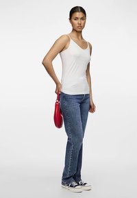 Hvit, ermfri tanktopp med tettsittende design, kombinert med blå jeans med rette ben og rød håndveske. Modellen står med avslappet holdning.