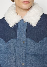 Gros plan sur une personne portant une veste en denim bleu avec un col en fourrure blanche et des boutons-pression en métal.
