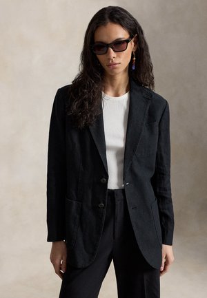 Hosszú, göndör hajú nő fekete napszemüvegben, fekete blézerben, fekete nadrágban, fehér bordázott felsőben és színes lógó fülbevalóval semleges háttér előtt.