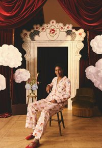 Růžový pyžamový set s hravým vzorem, který obsahuje směs barevných ilustrací. Dřevěná židle, červené texturované bačkůrky a květinové pozadí.