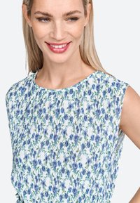 Ziedu raksta blūze ar ziliem un zaļiem ziediem uz balta fona, ar plisētu tekstūru un apaļu kaklu.