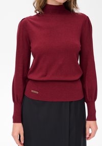 Kobieta w dopasowanym bordowym swetrze z długim rękawem i marszczonymi mankietami, w czarnej spódnicy, stojąca na jednolitym białym tle.