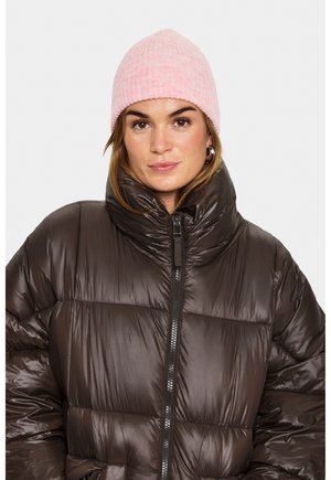 Kvindelig iført en skinnende mørkebrun pufferjakke med høj krave og en lyserød strikbeanie, der står fremadskuende med et neutralt udtryk.