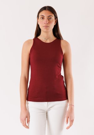 Donna con capelli lunghi e scuri che indossa un top senza maniche color bordeaux e pantaloni bianchi, su sfondo chiaro uniforme, rivolta verso l'osservatore.