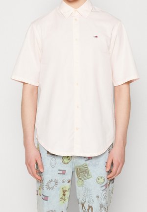 Mann trägt ein weißes, kurzärmeliges Hemd mit Knöpfen und kleinem Logo sowie hellblaue Jeans mit verschiedenen bunten Grafiken.